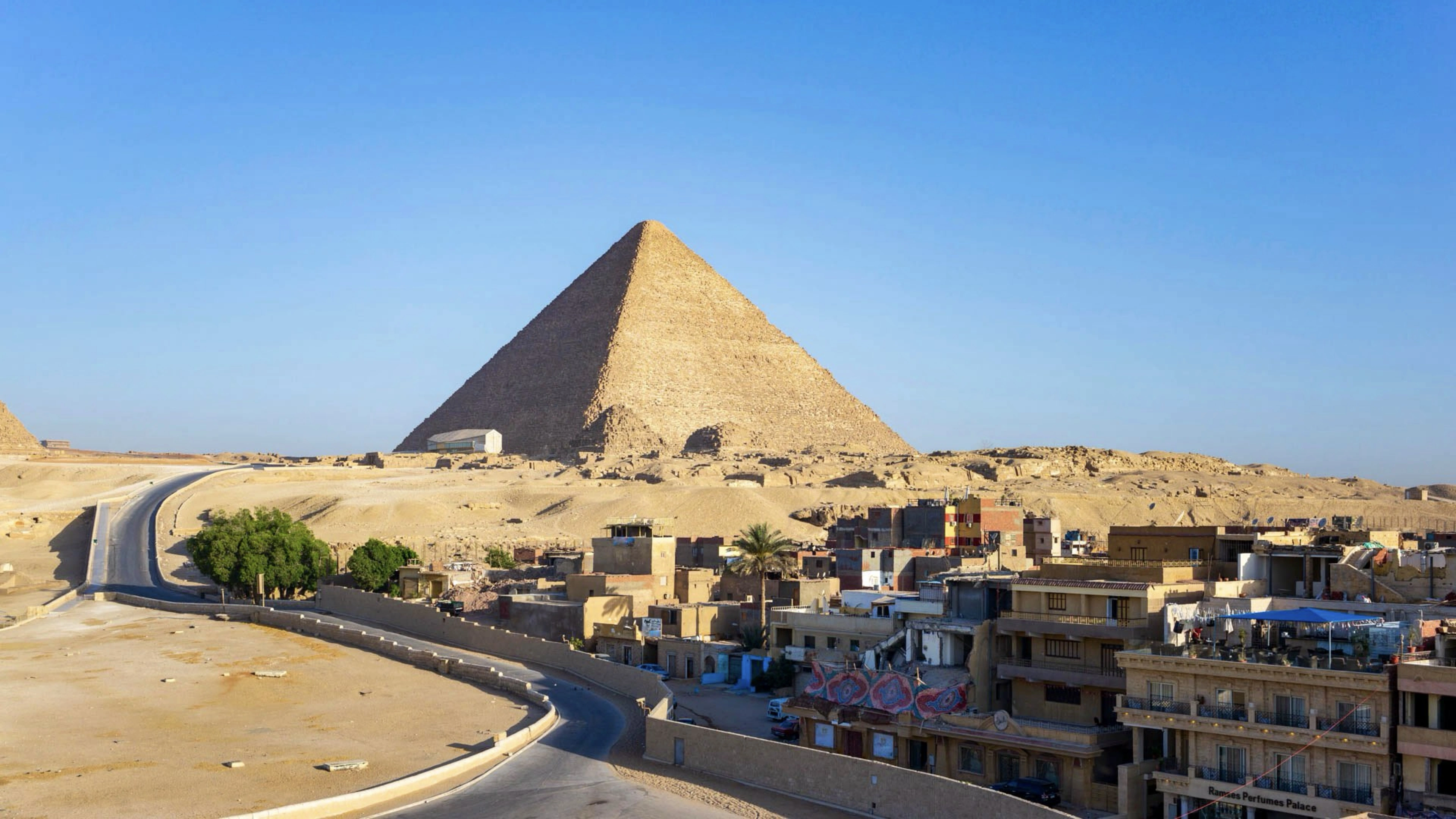 A view of the Great Pyramid of Cheops in Giza, Egypt, set against a clear blue sky. The foreground features a winding road leading towards the pyramid, with several buildings and small structures situated near the base of the pyramid. Photo by FlorentMartin_ via <a href='https://www.flickr.com/photos/yoyo0/49626979137/in/photolist-2iBneVk-2dxXwk8-RZ9gmP-2nN3fE8-2cRyz7X-2dr86UX-2gv1A1y-23RchDZ-SKNMUU-2iFBRf1-abMDxk-2dLgkSW-2mLjugE-2o9Nqa1-2ankvjA-2hGw2GG-yBNw1C-2kdBX4N-2oGuiNN-2pnZnEK-2d9bjTt-2iFBsHT-2bsYksX-7ULiJd-b1dQHk-b1dPjk-2exU8xE-2omRrZf-2ojvSzG-2dBPuBr-2phAYVU-2ikxp7X-2iRH3Nm-ZUxukP-2e2cUJx-8HpmPX-2bozDN5-FT3XkW-2kUKLTH-ZRRP17-X26pGM-2p4LPcL-2kqqUrm-2mLwX6p-2e2cUkM-jurjGS-2kzg4ws-AyfeJQ-nwUcy3-4ziFZn'>Flickr</a>