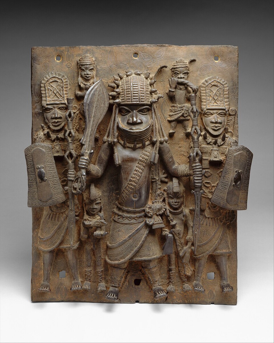 Bronze relief plaque from the Benin Kingdom depicting a central warrior figure in elaborate regalia, flanked by attendants and guards, all richly adorned with patterned armor and ceremonial weapons. Benin plaque with warriors and attendants (16th–17th century) via [Wikimedia](https://en.wikipedia.org/wiki/African_art#/media/File:Plaque-_Warrior_and_Attendants_MET_DT1231.jpg)