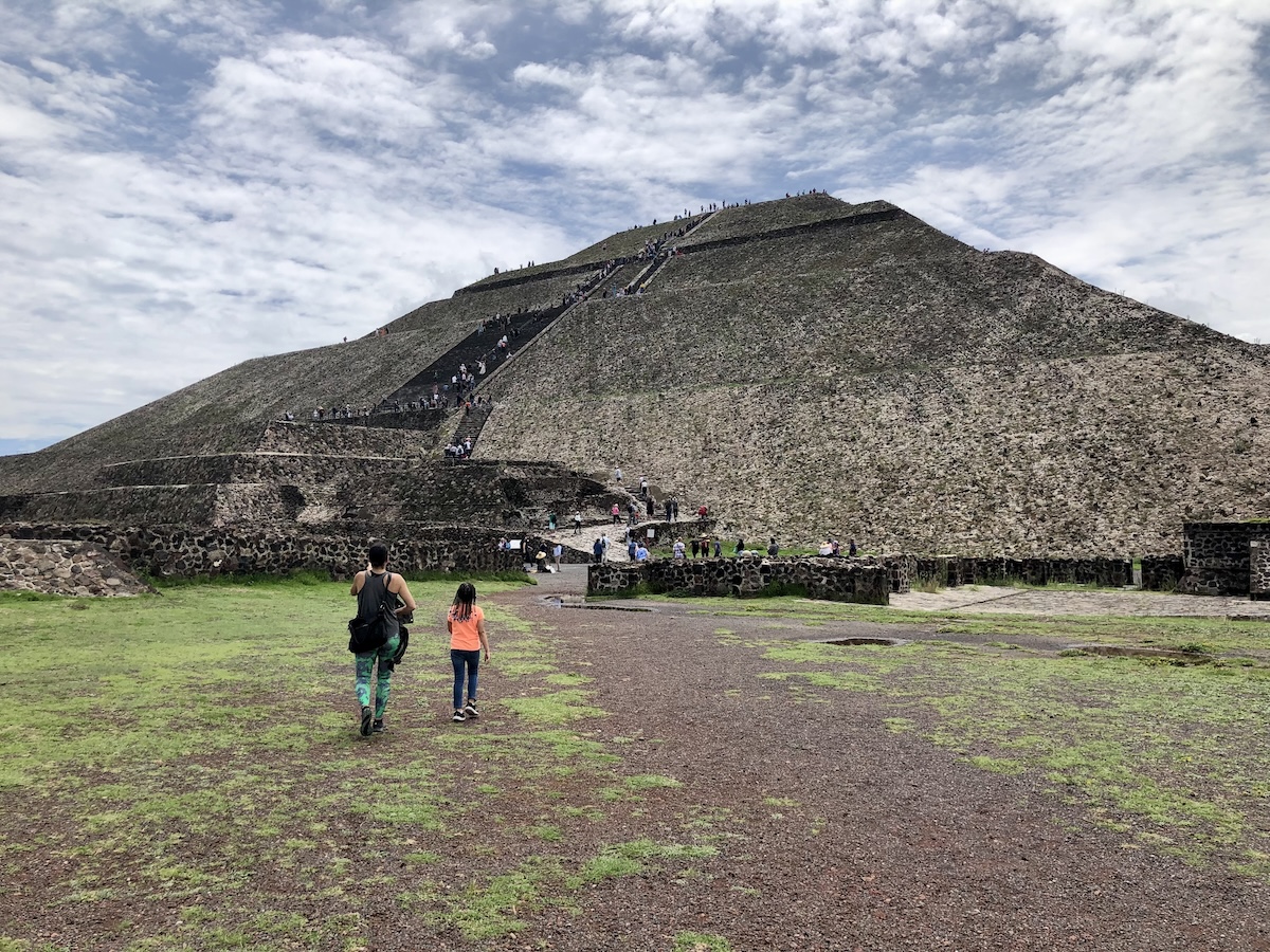 Visitors approach and climb the Pyramid of the Moon at Teotihuacan, a massive stepped stone structure set against a partly cloudy sky. My wife and daughter approaching the Pyramid of the Moon at Teotihuacan