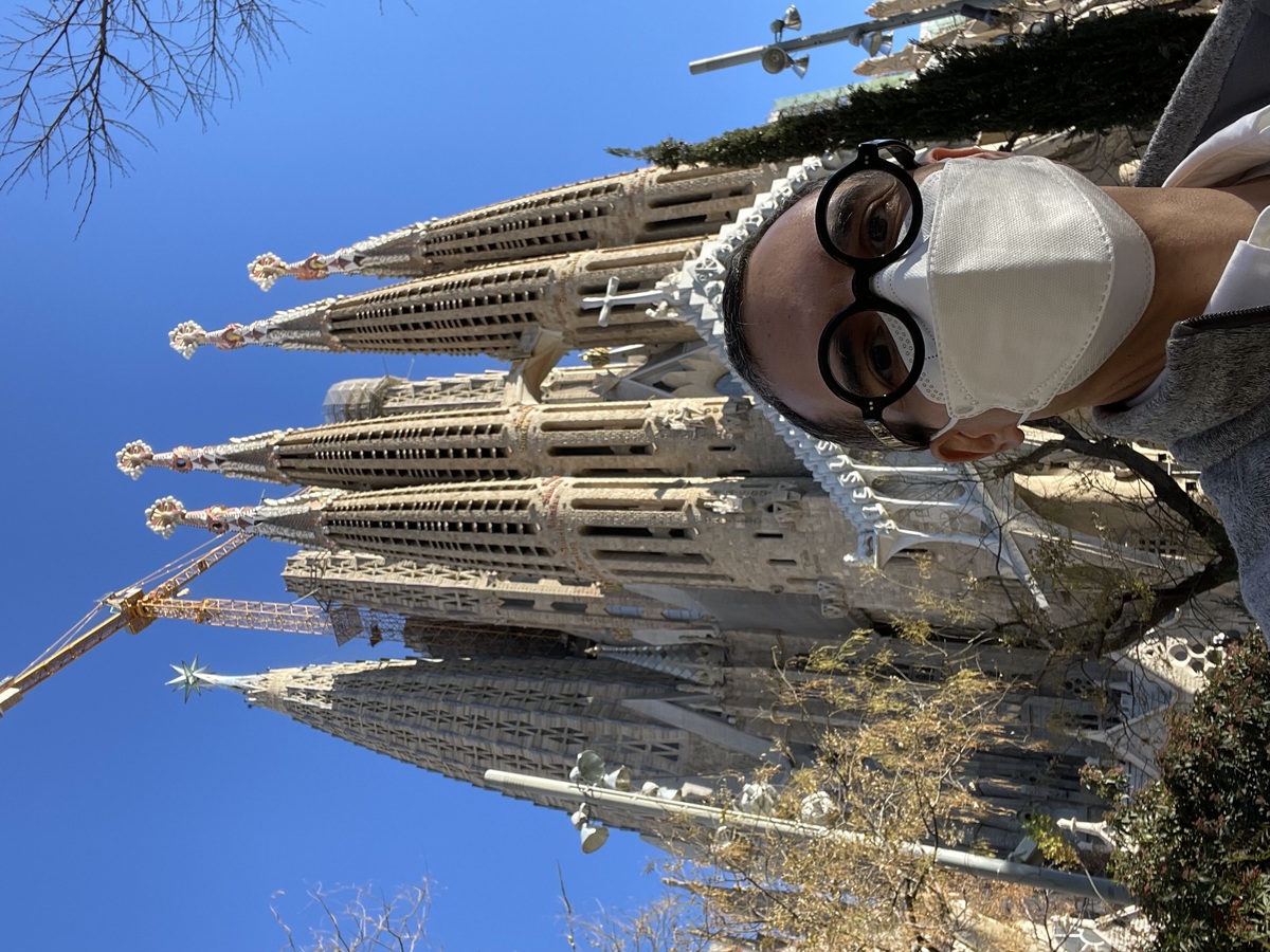 A person wearing glasses and a face mask stands in front of the Sagrada Familia in Barcelona, with its distinct spires and a construction crane visible. It’s about time I shared a selfie in the course.