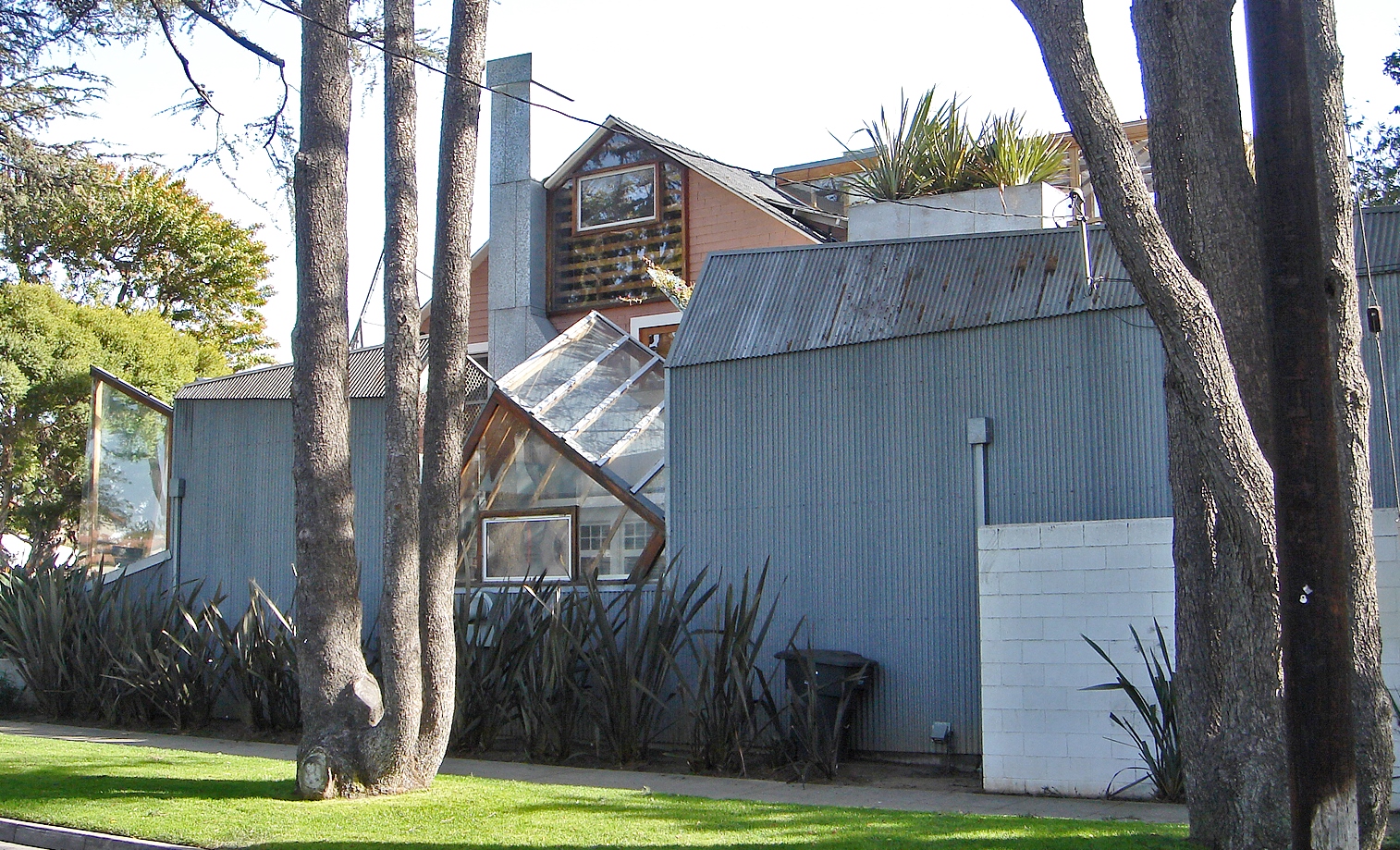 Corrugated metal building with unique geometric extensions and large windows, surrounded by trees and plants, under a clear sky. Photo of the Gehry Residence in Santa Monica by IK's World Trip via [Wikimedia](https://commons.wikimedia.org/w/index.php?curid=13364513)